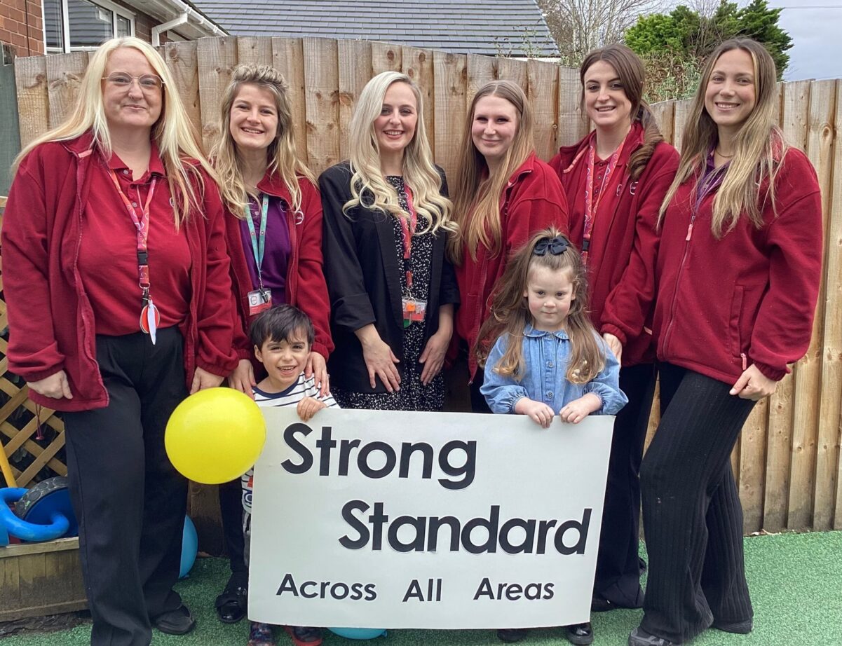 Team members and children from Partou Sunny Day Day Nursery & Pre‑school in Ellesmere Port stand outdoors holding a sign reading “Strong Standard Across All Areas” following their Ofsted inspection