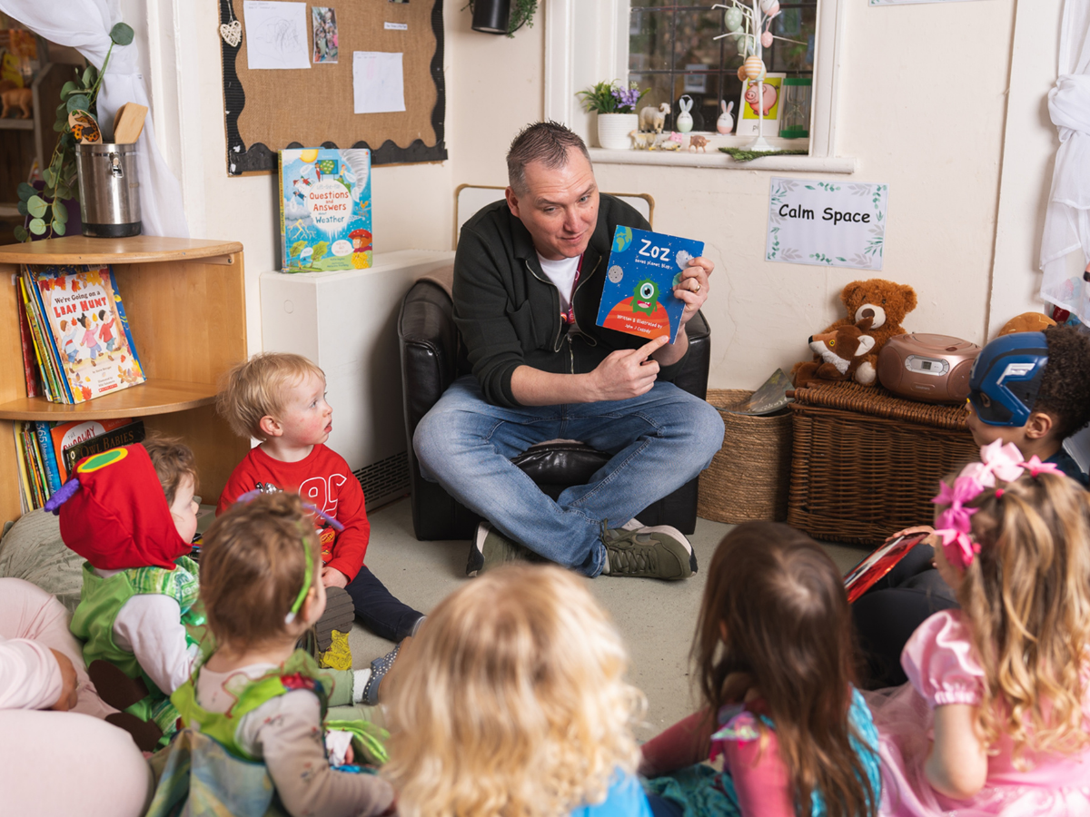 Children’s author John Cassidy sits reading a book aloud to a small group of young children during a storytime session at Partou Cherubs Day Nursery & Pre‑school in Sale.