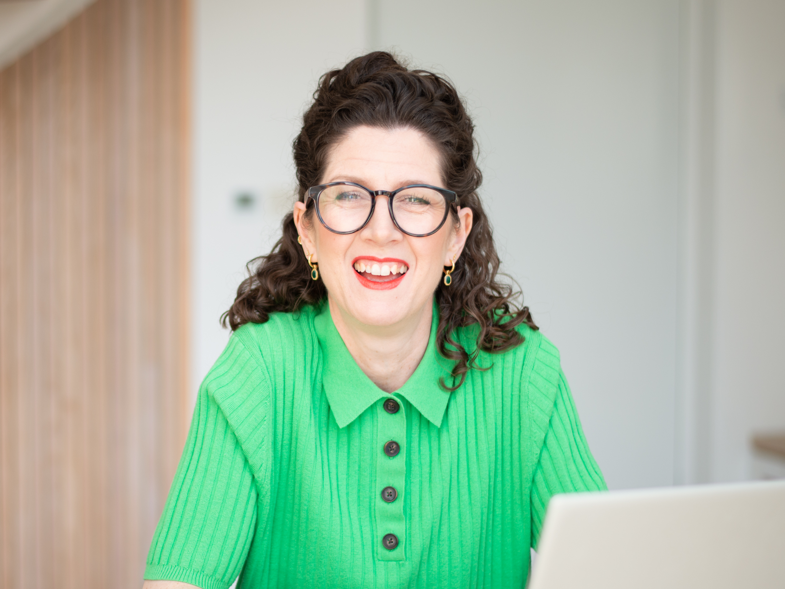 Laura Matthews, Early Years Nutritionist for Partou, sitting indoors in a bright green collared knit top, wearing round glasses and gold hoop earrings as she works on a light‑coloured laptop in a modern, softly lit setting.
