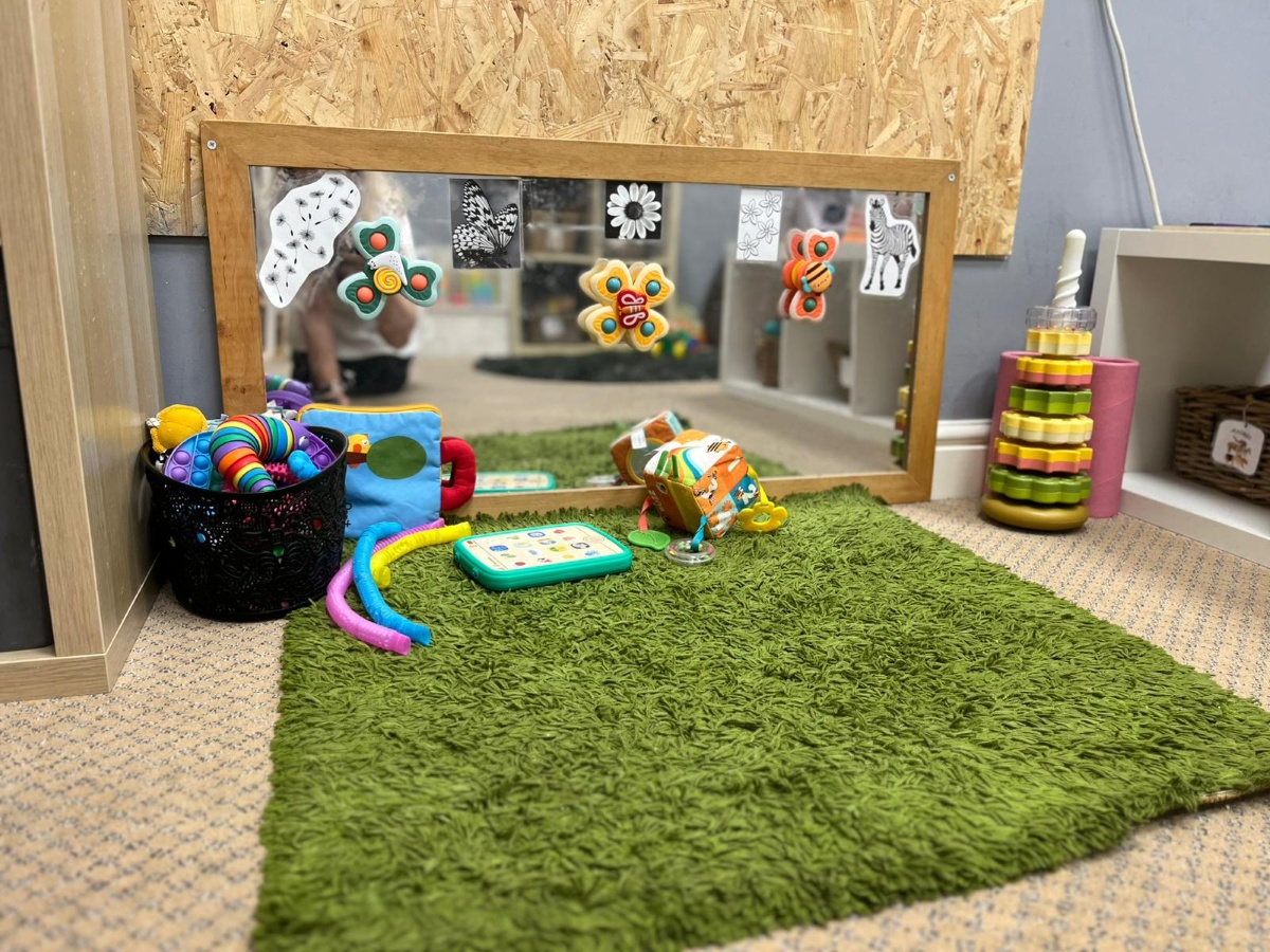 A cosy sensory play space at Partou Chorley Day Nursery & Pre‑School, featuring a green shaggy rug, a low wooden‑framed mirror decorated with colourful nature stickers, and a selection of baby toys including soft cubes, rattles and flexible tubes, with nearby shelves and baskets offering additional sensory resources for early exploration.