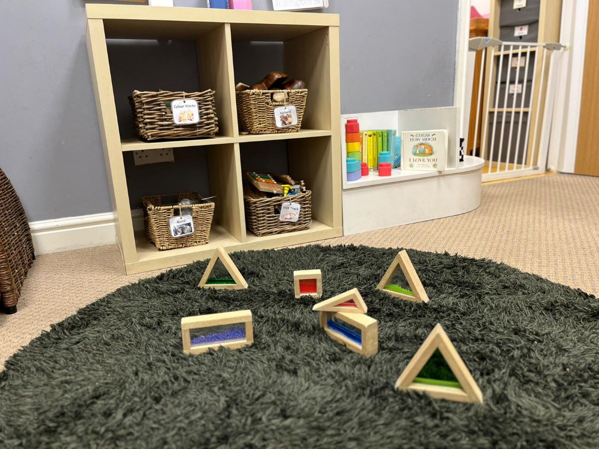 A neatly organised learning space at Partou Chorley Day Nursery & Pre‑School, featuring a dark green circular rug with colourful wooden sensory blocks, a wooden shelving unit with labelled wicker baskets, and a small white display shelf holding stacking toys and picture books, creating an inviting environment for early exploration and play.