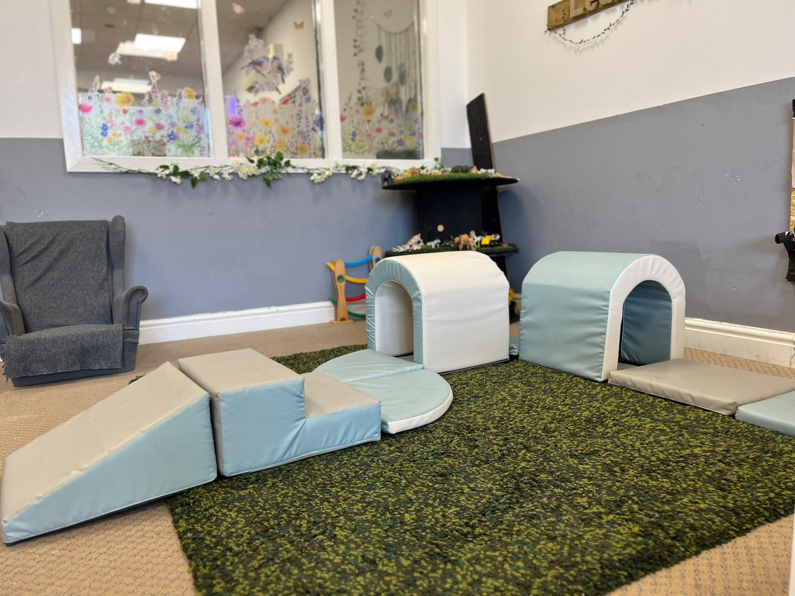 A neatly organised learning space at Partou Chorley Day Nursery & Pre‑School, featuring a dark green circular rug with colourful wooden sensory blocks, a wooden shelving unit with labelled wicker baskets, and a small white display shelf holding stacking toys and picture books, creating an inviting environment for early exploration and play.