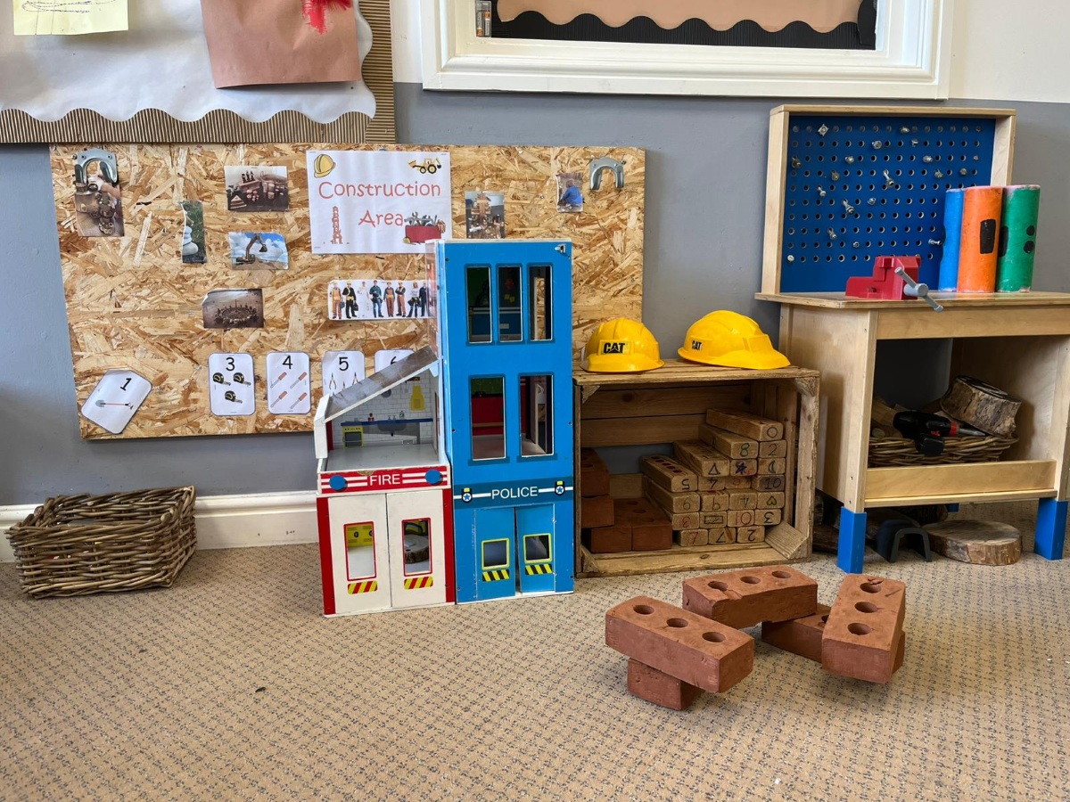 A construction‑themed play area at Partou Chorley Day Nursery & Pre‑School, featuring a wall display with construction photos and number cards, a toy building set labelled for fire and police play, yellow CAT helmets on a wooden shelf, numbered wooden blocks, realistic toy bricks on the floor and a small workbench with colourful tools to support imaginative building and role‑play.
