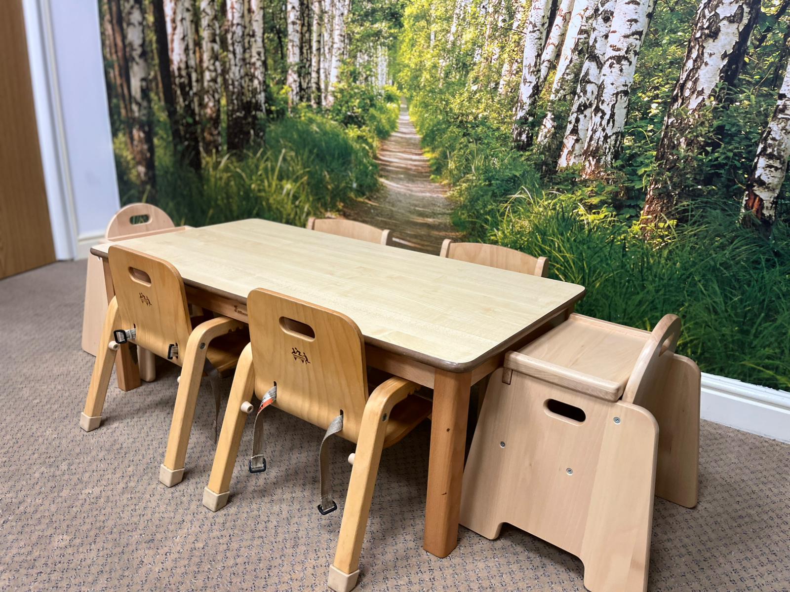 A wooden table surrounded by six child‑sized chairs at Partou Chorley Day Nursery & Pre‑School, set against a large forest‑themed wall mural featuring tall birch trees and a leafy woodland path, creating a calming, nature‑inspired space for group play and learning.