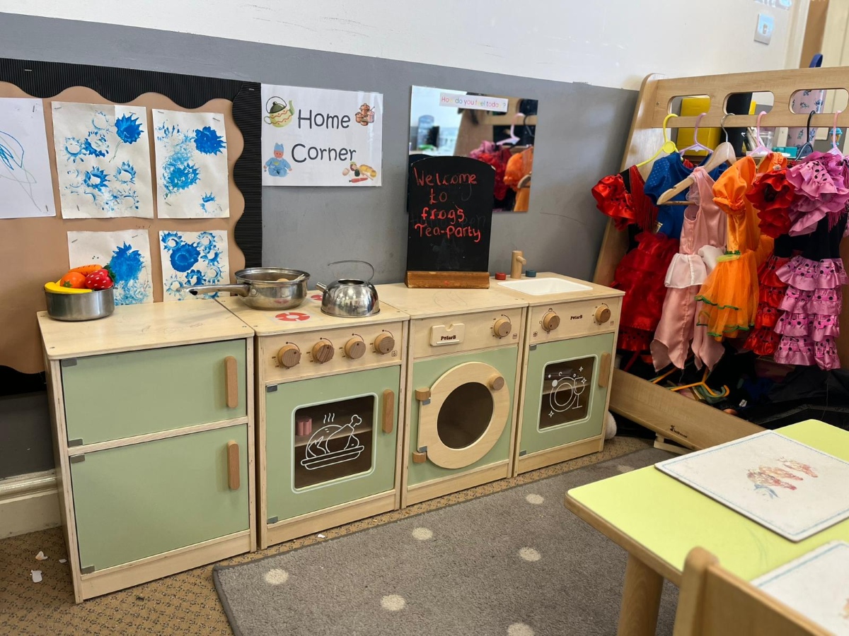 A colourful home‑corner play area at Partou Chorley Day Nursery & Pre‑School, featuring a wooden play kitchen with green and beige units, toy pots and play food, a rack of bright dressing‑up clothes and wall displays including a ‘Home Corner’ sign, children’s artwork and a chalkboard reading ‘Welcome to Frogs Tea‑Party’, creating a vibrant space for imaginative role‑play.