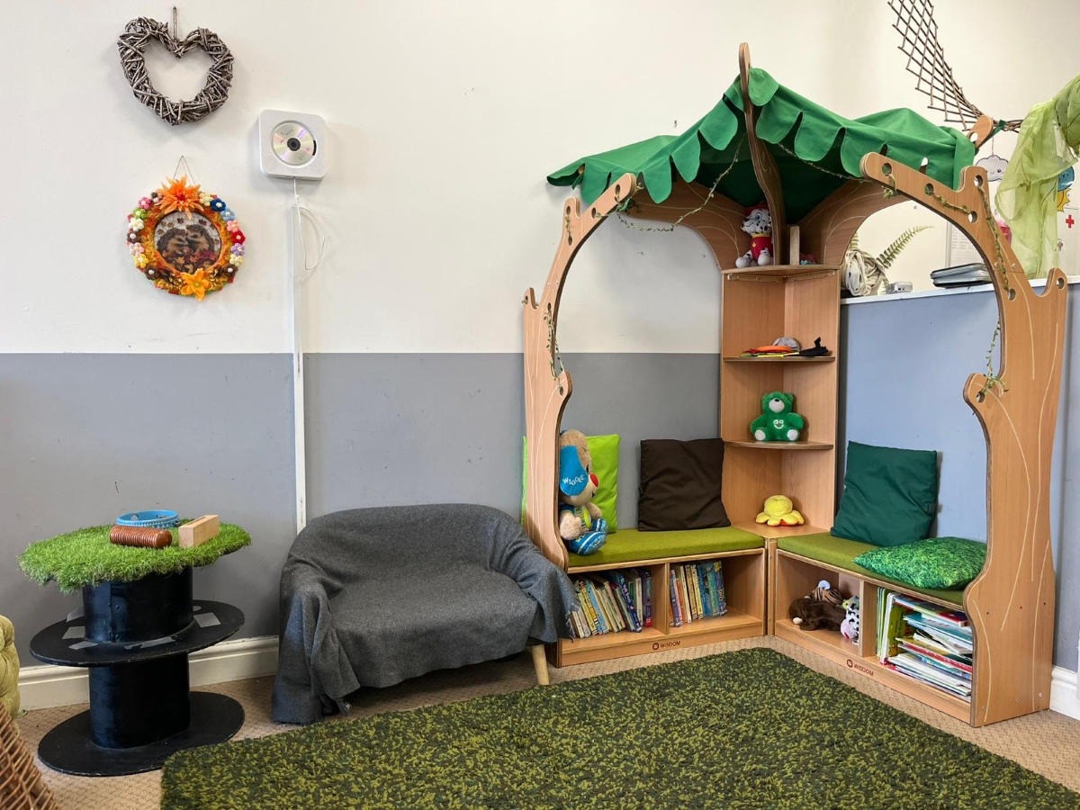 A cosy woodland‑themed reading nook at Partou Chorley Day Nursery & Pre‑School, featuring a wooden tree‑shaped frame with a green leaf canopy, shelves of books and soft toys, green and brown cushions, a small armchair with a blanket and a round grass‑topped table with wooden blocks, creating a calm, nature‑inspired space for quiet play and stories.