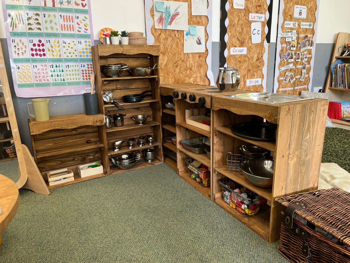 A wooden role‑play kitchen area at Partou Chorley Day Nursery & Pre‑School, featuring shelves of metal pots, pans and jugs, a built‑in sink and stovetop, and wall displays showing numbers, letters, colours and children’s artwork, creating a practical life‑inspired space for imaginative play and early learning.