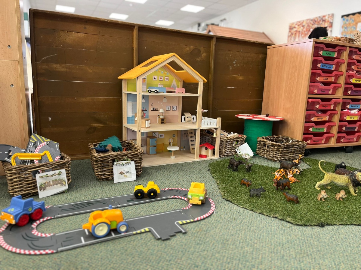 A small‑world play area at Partou Chorley Day Nursery & Pre‑School, featuring a wooden dollhouse with miniature furniture, wicker baskets of dinosaurs and road pieces, a grassy play mat with toy animals, and a road track with small vehicles, set beside a wooden storage unit with labelled red drawers to support imaginative storytelling and early learning.