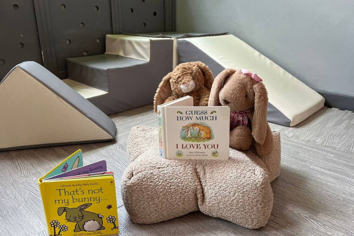 Two soft plush bunnies sitting on a beige cushion at Partou Heskin Day Nursery & Pre‑School, positioned as if reading children’s books including “Guess How Much I Love You” and a touch‑and‑feel bunny story, with a grey soft‑play ramp and steps in the background creating a calm, inviting reading and play space.