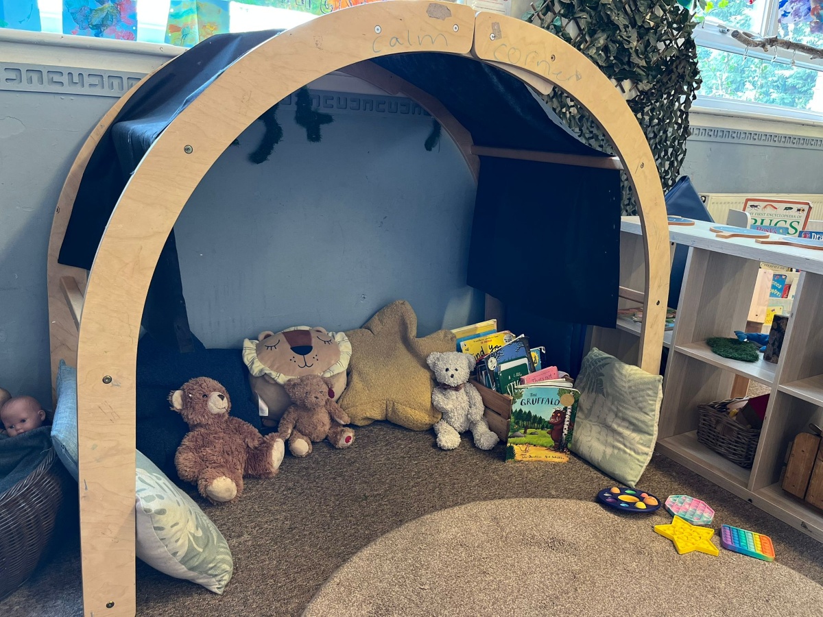 A cosy ‘calm corner’ at Partou Heskin Day Nursery & Pre‑School, framed by a wooden arch and filled with soft teddy bears, star‑shaped cushions, picture books and colourful sensory toys, creating a comforting space for children to relax and regulate, surrounded by shelves of baskets and a bright classroom window display.