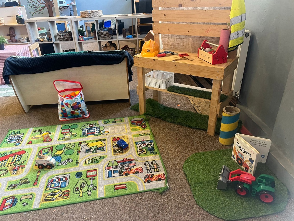 A vibrant small‑world play area at Partou Heskin Day Nursery & Pre‑School, featuring a colourful city‑themed play mat with toy cars and trucks, a wooden workbench with pretend tools and a yellow safety vest, wicker baskets on artificial grass, and a toy tractor beside a children’s book, all set within a bright, well‑organised classroom space.