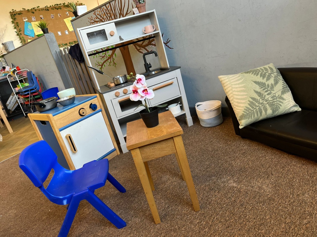 A bright and welcoming play area at Partou Heskin Day Nursery & Pre‑School, featuring a small wooden table with a potted orchid, a blue child‑sized chair, a wooden toy kitchen with pots and cups, and a cosy black sofa with a fern‑pattern cushion, set against shelves of play resources and a colourful classroom display.