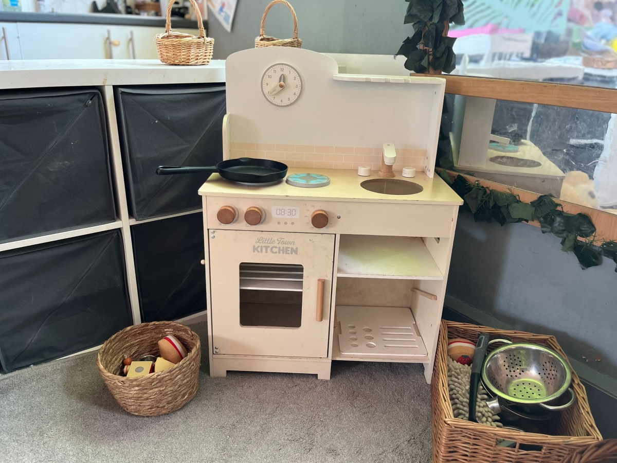 A cream wooden play kitchen at Partou Heskin Day Nursery & Pre‑School, complete with a toy stove, sink and oven, surrounded by wicker baskets filled with pretend food and metal utensils, set beneath a mirror framed with greenery and soft storage baskets in a bright, child‑friendly play space.