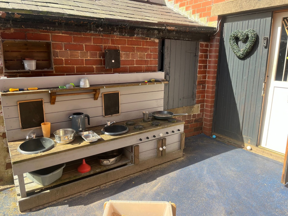 A wooden outdoor play kitchen at Partou Heskin Day Nursery & Pre‑School, set against a red‑brick wall, with metal bowls, a kettle and colourful utensils arranged for imaginative play, alongside two small chalkboards and a door decorated with a green heart‑shaped wreath on a bright blue play surface.