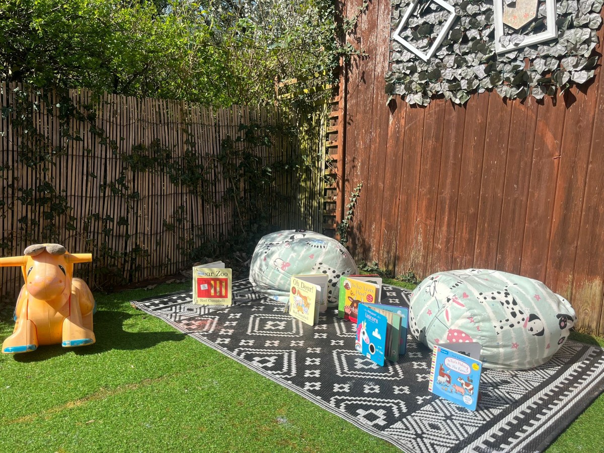 A cosy outdoor reading nook at Partou Heskin Day Nursery & Pre‑School, with a black‑and‑white patterned rug, two light‑coloured beanbags, a selection of bright children’s books including animal‑themed titles, and an orange ride‑on toy set against a wooden fence decorated with mirrors and greenery on soft artificial grass.