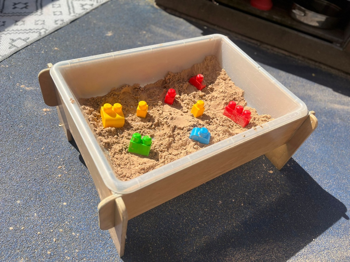 A sunlit outdoor sensory tray at Partou Heskin Day Nursery & Pre‑School, filled with soft light‑brown sand and scattered with bright red, yellow, green and blue building bricks, set within a wooden frame on a blue play surface to encourage creative, hands‑on exploration.