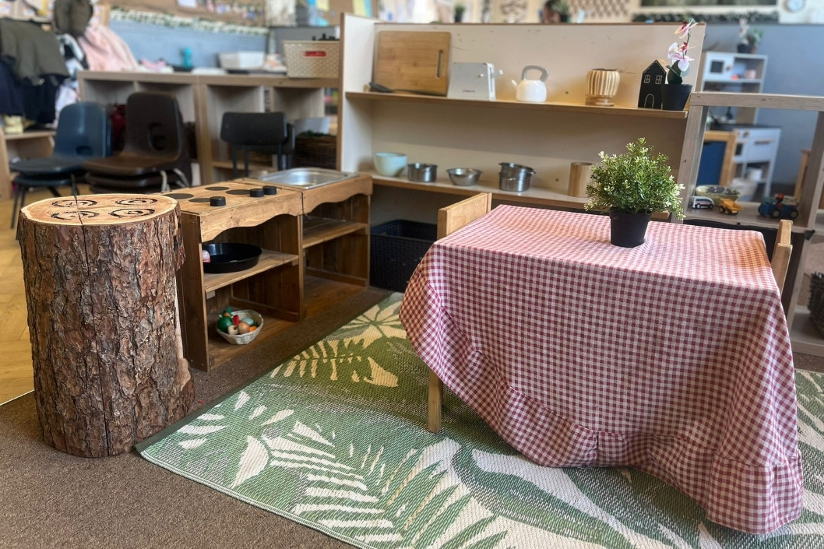 A cosy indoor role‑play area at Partou Heskin Day Nursery & Pre‑School, featuring a wooden play kitchen with pots, pans and bowls, a small table with a red‑and‑white check tablecloth and potted plant, a natural wooden log seat and a green leaf‑pattern rug, surrounded by shelves of imaginative play resources and children’s coats.
