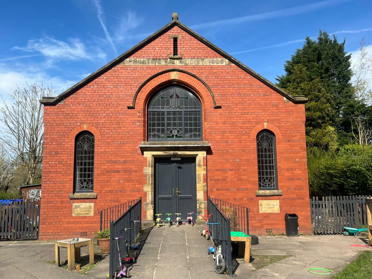 The historic red‑brick 1905 Primitive Methodist building now home to Partou Heskin Day Nursery & Pre‑School, with colourful children’s scooters and trikes arranged on the paved outdoor area, framed by wooden fencing, trees and a bright blue sky.