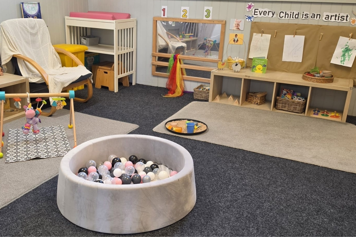 A calm, creative play space at Partou Parbold Day Nursery & Pre‑School in Wigan, featuring a soft circular ball pit with pastel balls, a wooden baby gym on a patterned mat, a rocking chair with a cosy blanket, a changing area, a large mirror with colourful scarves, and a wooden shelf filled with baskets, books and art materials beneath a display reading ‘Every Child is an artist’.