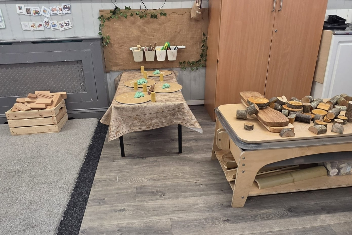 A nature‑inspired creative play area at Partou Parbold Day Nursery & Pre‑School in Wigan, featuring wooden blocks, log slices, natural loose parts and crafting tools arranged on rustic tables with hanging pots and an ivy‑trimmed backdrop, encouraging hands‑on exploration, fine‑motor development and imaginative learning.