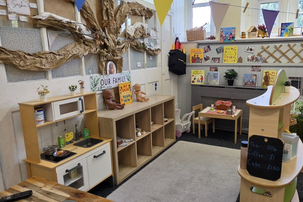 A welcoming role‑play area at Partou Parbold Day Nursery & Pre‑School in Wigan, featuring a wooden play kitchen, a miniature table and chairs, shelves filled with toy dishes and dolls, and a large paper ‘Our Family’ display tree, surrounded by diverse children’s books, potted plants and colourful bunting to create a homely, imaginative learning space.