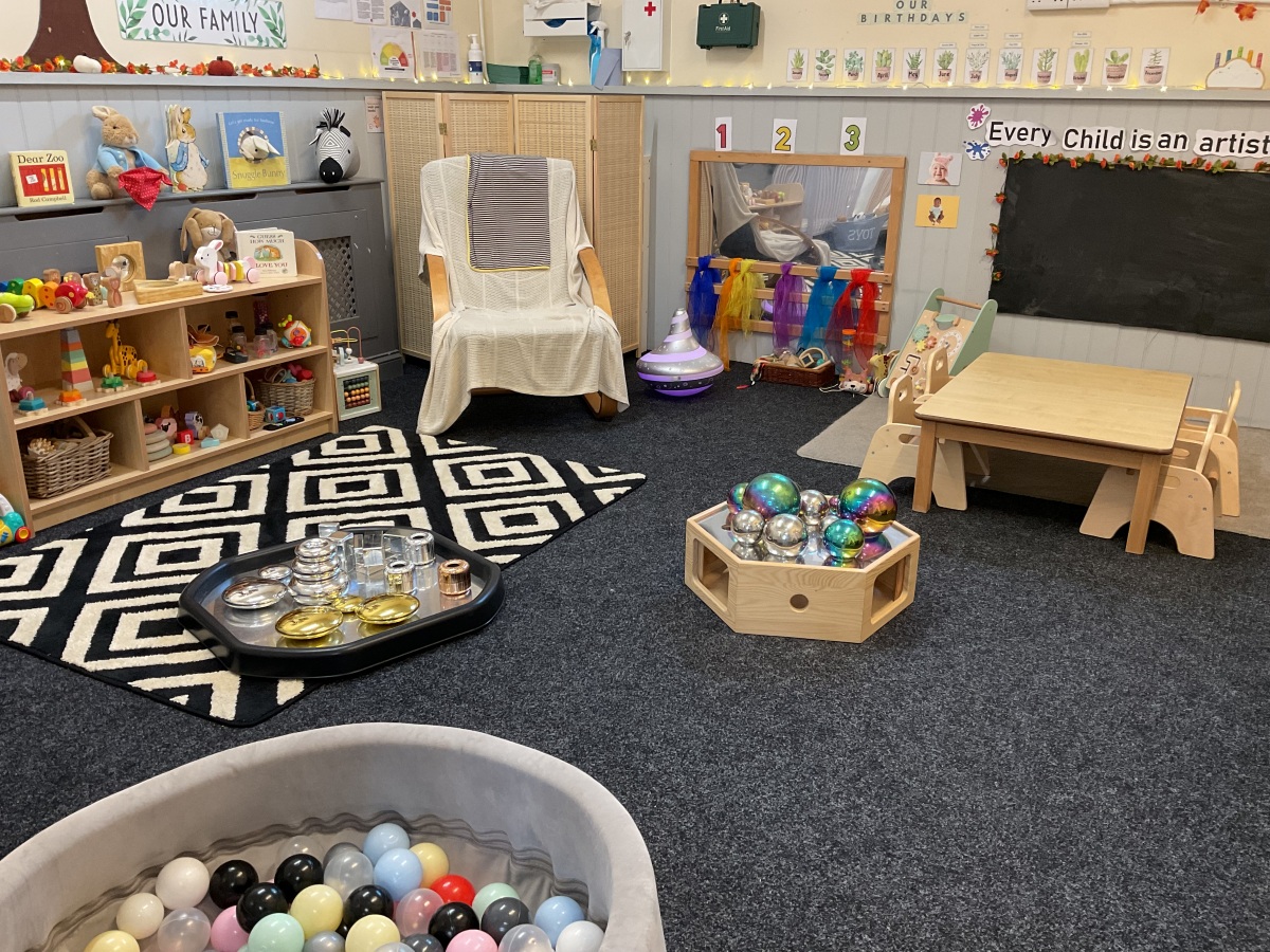 Welcoming baby room at Partou Parbold Day Nursery & Pre-school with natural wood tables and chairs, soft grey flooring, a black chalkboard wall, cosy sensory play areas and colourful learning resources in red, blue, green, orange and yellow. Designed to support babies