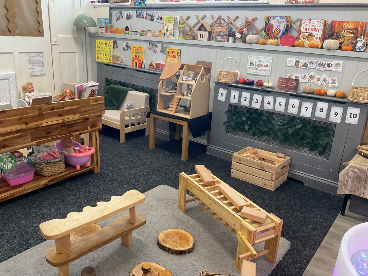 Inspiring learning environment at Partou Parbold Day Nursery & Pre-school combining mathematical learning, movement and imaginative play. Natural wood balance equipment and construction resources sit alongside number displays, while soft grey flooring, sage green foliage panels, cream furnishings and autumn decorations in warm orange, rust and pumpkin tones create a stimulating space for problem-solving, physical development and creative exploration.