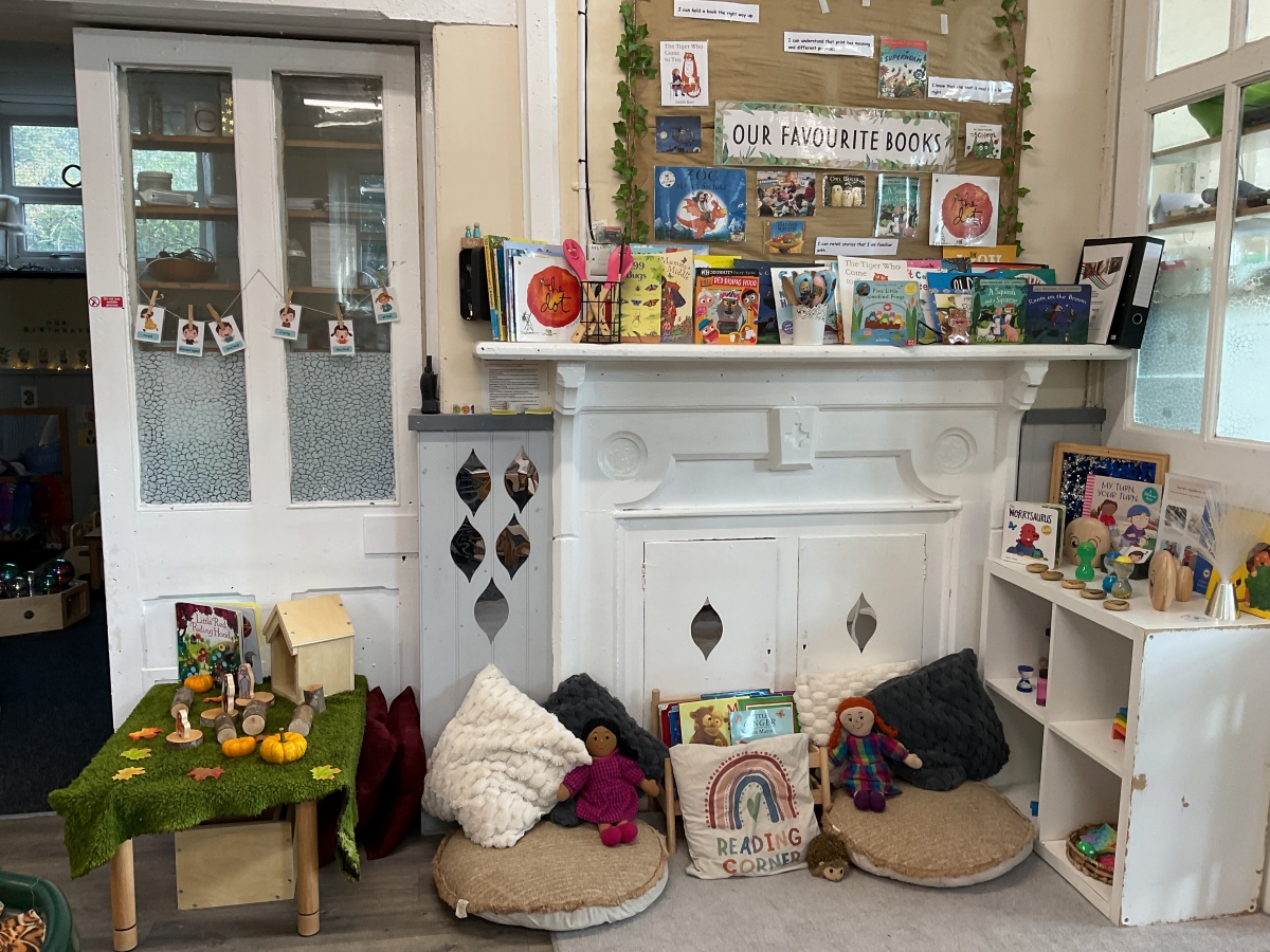 Cosy reading corner at Partou Parbold Day Nursery & Pre-school featuring a traditional white fireplace, natural wood bookshelves, soft cream and charcoal cushions, woven floor cushions and a carefully curated children