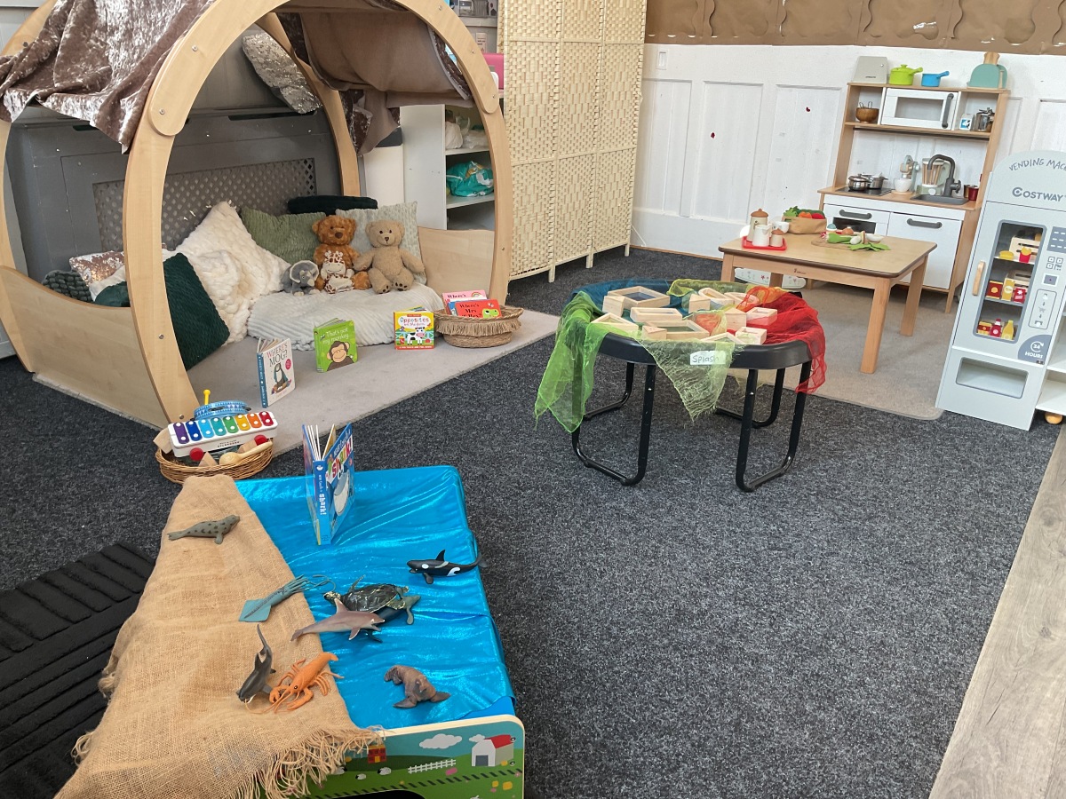 Inviting small world and sensory play area at Partou Parbold Day Nursery & Pre-school, featuring a natural wooden dolls’ house, cosy reading den, soft play shapes in grey and cream, and hands-on exploration resources. Bright blue sensory fabric, colourful translucent scarves in red, green and yellow, and natural wood construction materials create an engaging early years environment that encourages imaginative play, communication skills and child-led learning.
