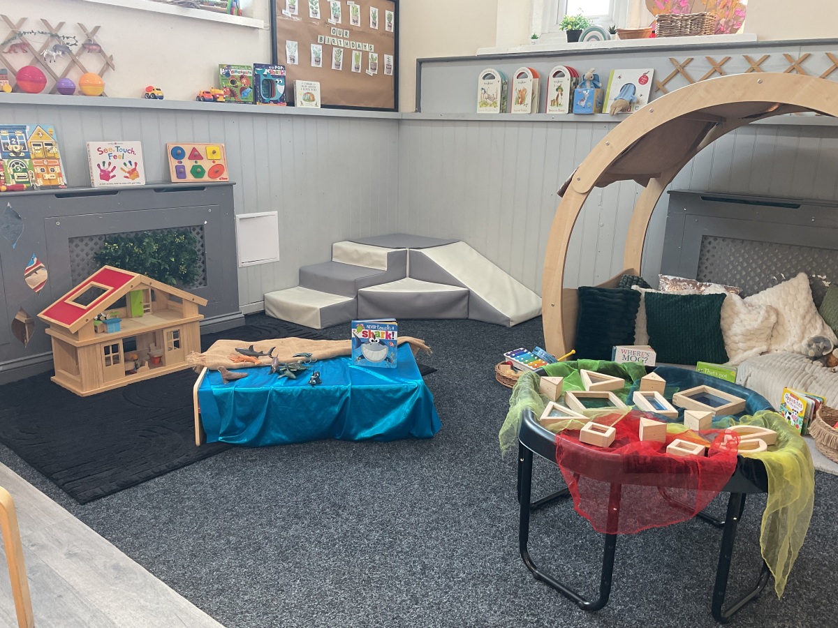Creative learning space at Partou Parbold Day Nursery & Pre-school combining sensory exploration, role play and early literacy opportunities. A beautiful wooden play den with soft cream, sage green and charcoal cushions sits alongside a role-play kitchen, ocean-themed small world activity with vibrant blue fabric, and sensory resources draped in red and green sheer materials. The warm neutral palette, natural wood furnishings and colourful learning resources support curiosity, problem-solving and learning through play in an inspiring nursery environment.