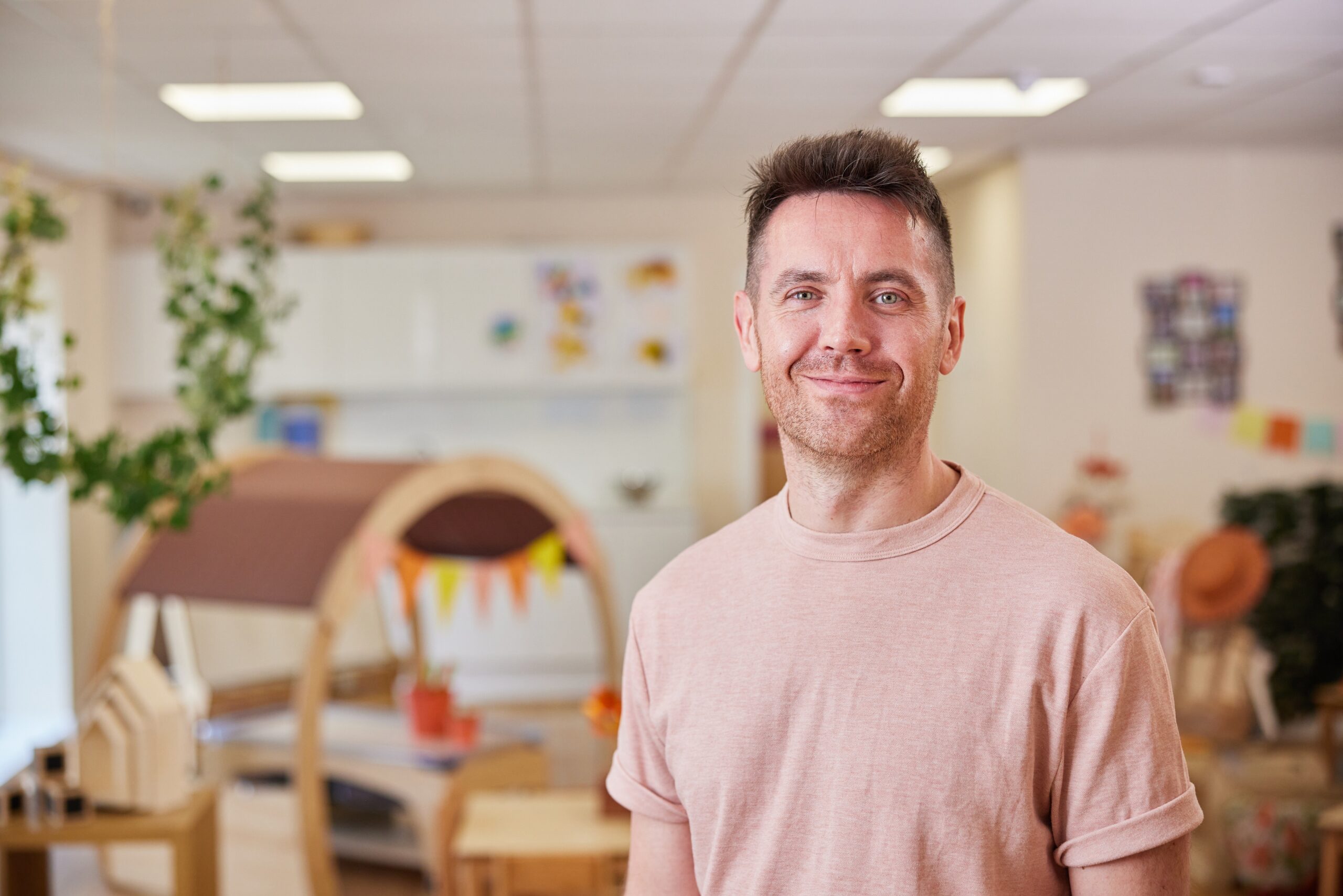 Head-and-shoulders photograph of a person wearing a light pink T‑shirt, standing in a nursery room with wooden play equipment, plants and soft furnishings visible in the background.