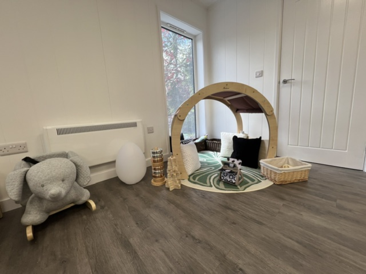 Bright, newly refurbished baby room at Partou Field View nursery in Alfreton, featuring a wooden activity arch, soft play cushions, neutral décor, sensory toys, wicker storage basket and a plush elephant rocker on wood flooring near a large window.