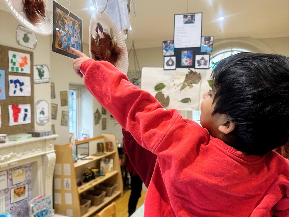 A child in a classroom points up at a display of artwork and craft pieces hanging from the ceiling and walls, including leaf collages, paintings and photographs. The room features educational displays, low shelving with toys and materials, and a decorated fireplace, creating a creative and engaging learning environment.