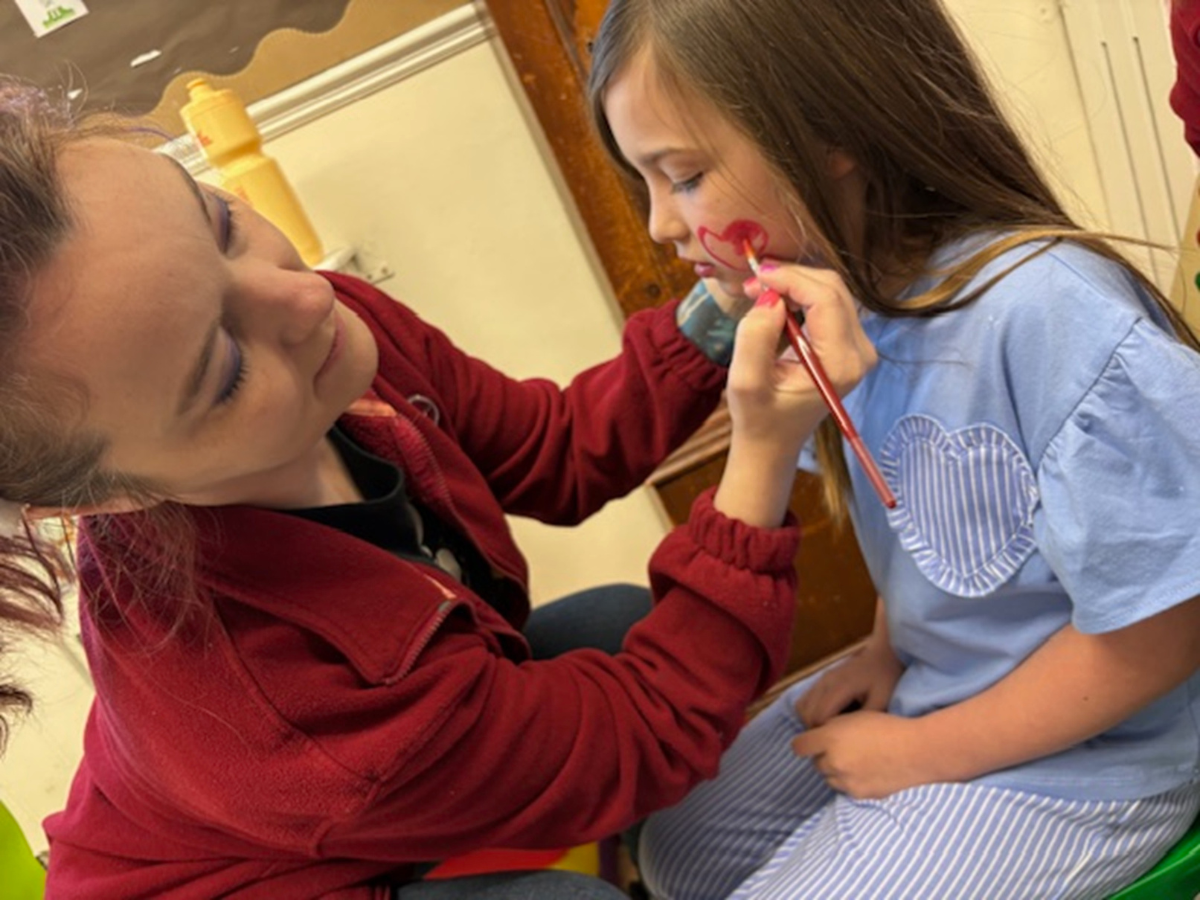 A childcare practitioner kneels beside a young child indoors, carefully painting a red heart shape on the child’s cheek with a small brush. The child sits on a low chair wearing a light blue outfit with a heart design, while classroom items and furniture are visible in the softly lit background.