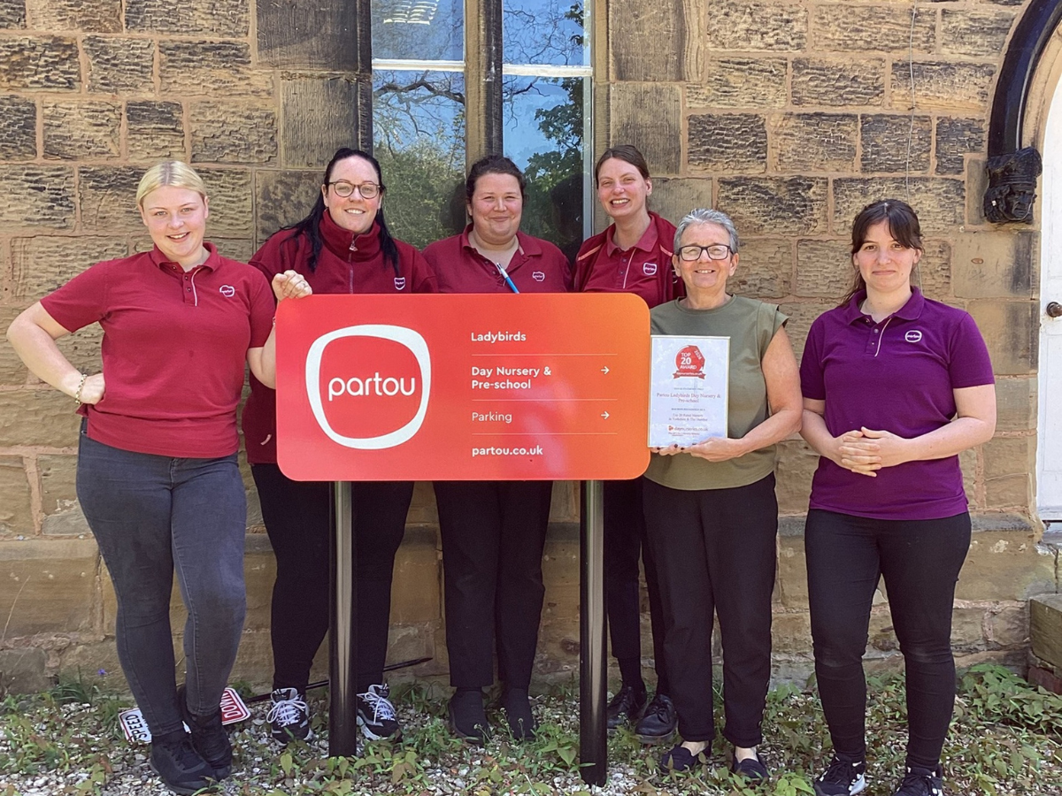 A group of nursery staff stand outside a stone building beside a red Partou Ladybirds Day Nursery & Pre-School sign, with one person holding an award plaque, marking a recognition achievement.