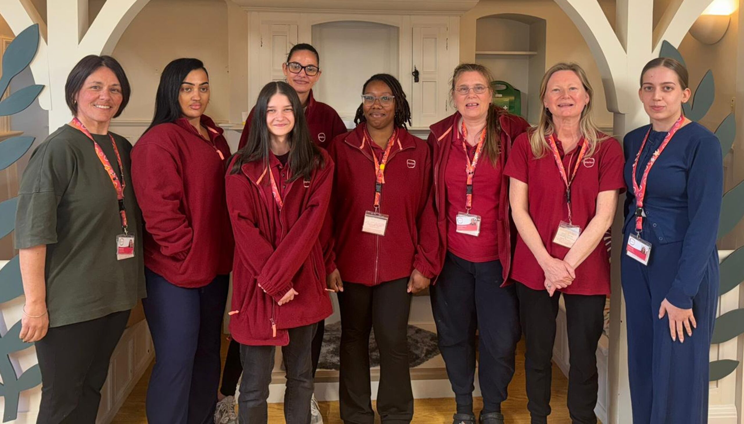A group of eight Partou Gingerbread House team members standing together indoors, wearing matching maroon uniforms and name badges, smiling for a team photo against a light, modern nursery backdrop.