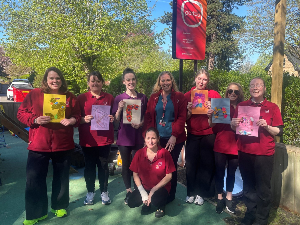 A group of childcare staff stand outdoors in a garden play area, holding up colourful children’s artwork. Behind them is a red Partou “Safehands Day Nursery & Pre-school” sign, with trees, greenery, and playground equipment visible in the sunny background.