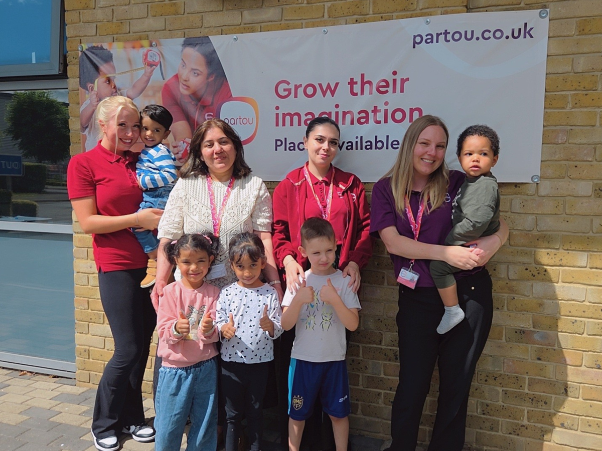 Group of childcare staff and young children standing outside in front of a brick wall. Several adults wear nursery uniforms and lanyards, and some are holding toddlers. Three children stand at the front giving thumbs up gestures. Bright daylight illuminates the scene.