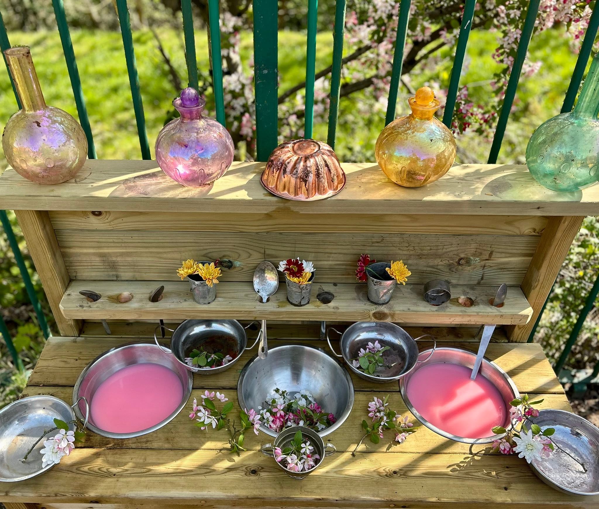A wooden mud kitchen outside in dappled sunlight, with iridescent pink, amber and pale green glass bottles and a copper jelly mould along the top shelf, small galvanised buckets of yellow, red and white flowers on the middle shelf, and silver metal pans below filled with bright pink water and scattered pink apple blossom, a calming water play setup for keeping children cool in hot weather.