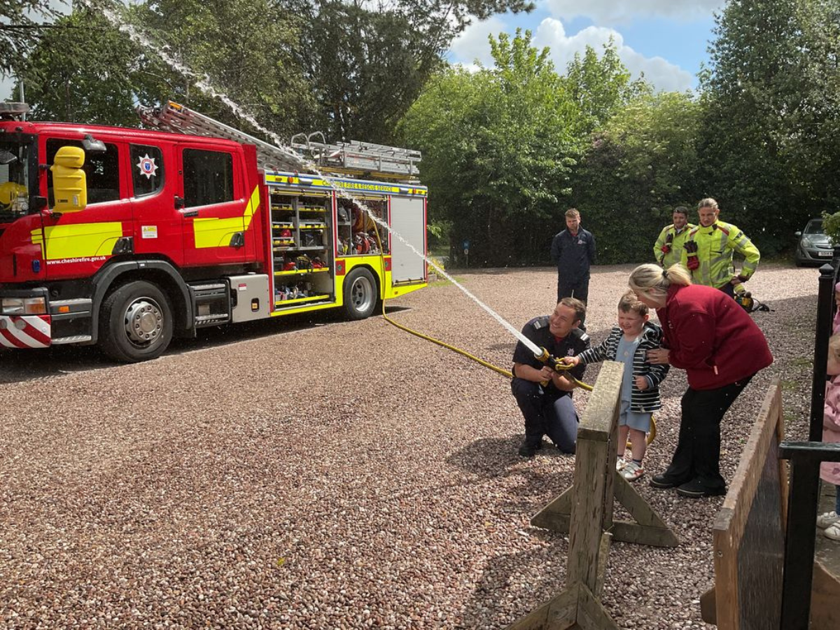 A fire engine from Cheshire Fire and Rescue Service visits Partou Kids Corner Day Nursery & Pre-school in Sandbach. A firefighter helps a child hold a fire hose, spraying water across an outdoor gravel area, while nursery staff and other firefighters watch. Trees and greenery surround the scene on a bright day.