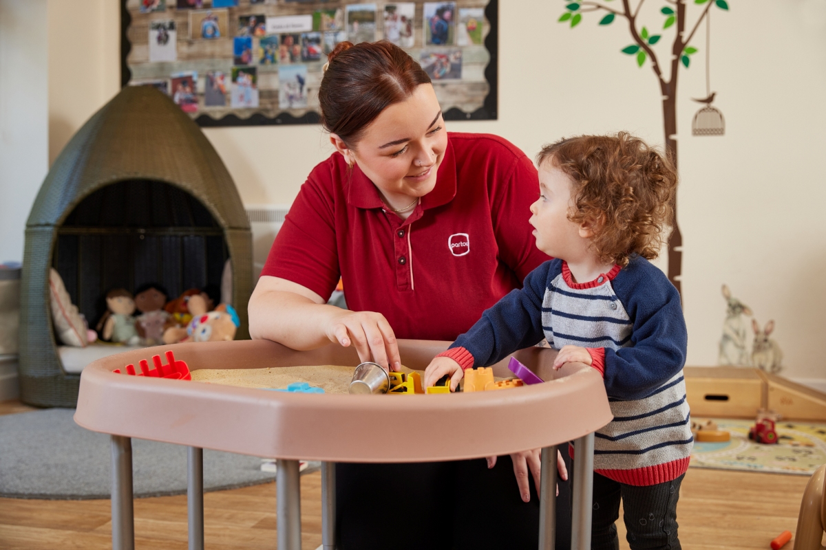 A Partou practitioner in a red polo shirt crouches beside a toddler at a blush-pink sand tray, making eye contact and smiling warmly as the child explores the sand with yellow and orange toy diggers. The practitioner