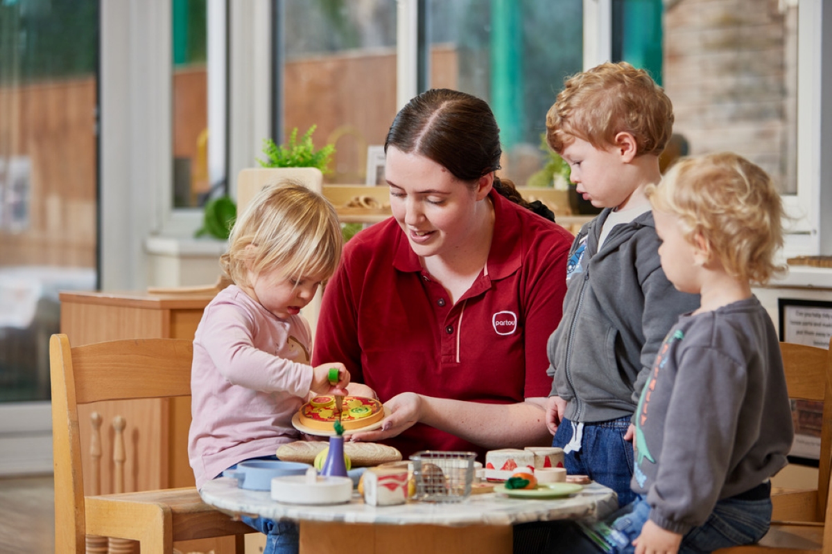 A Partou practitioner in a red polo shirt sits at a low table with three young children, holding a wooden toy pizza as a toddler reaches in to engage with it. The practitioner is leaning in attentively, smiling and following the children