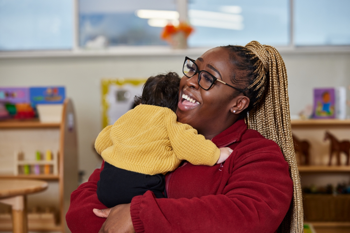 A Partou nursery key person in a red fleece holding and cuddling a young child in a yellow jumper, smiling warmly in a nursery classroom setting, supporting separation anxiety through secure, caring relationships.