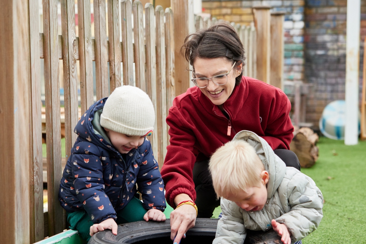 A Partou practitioner in a red fleece crouches down in an outdoor play area with two toddlers, all three focused on exploring a tyre together. The practitioner is smiling broadly, pointing into the tyre and fully absorbed in the moment alongside the children. Both children, one in a navy fox-print puffer jacket and cream beanie and one in a silver-grey hooded coat, are leaning in with curiosity. The image captures the kind of warm, present, and unhurried adult attention that helps children feel safe and confident to explore.