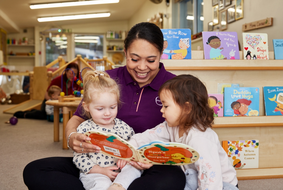 12:04Claude responded: A Partou practitioner in a purple polo shirt sits cross-legged on the floor of a nursery book corner, with two toddlers nestled close beside her as she holds o…A Partou practitioner in a purple polo shirt sits cross-legged on the floor of a nursery book corner, with two toddlers nestled close beside her as she holds open a board book. She is smiling warmly as all three look at the pages together. One child, in a white leopard-print top, leans in from her lap while the other, in a white floral top, reaches towards the book with interest. The relaxed, floor-level setting and the children