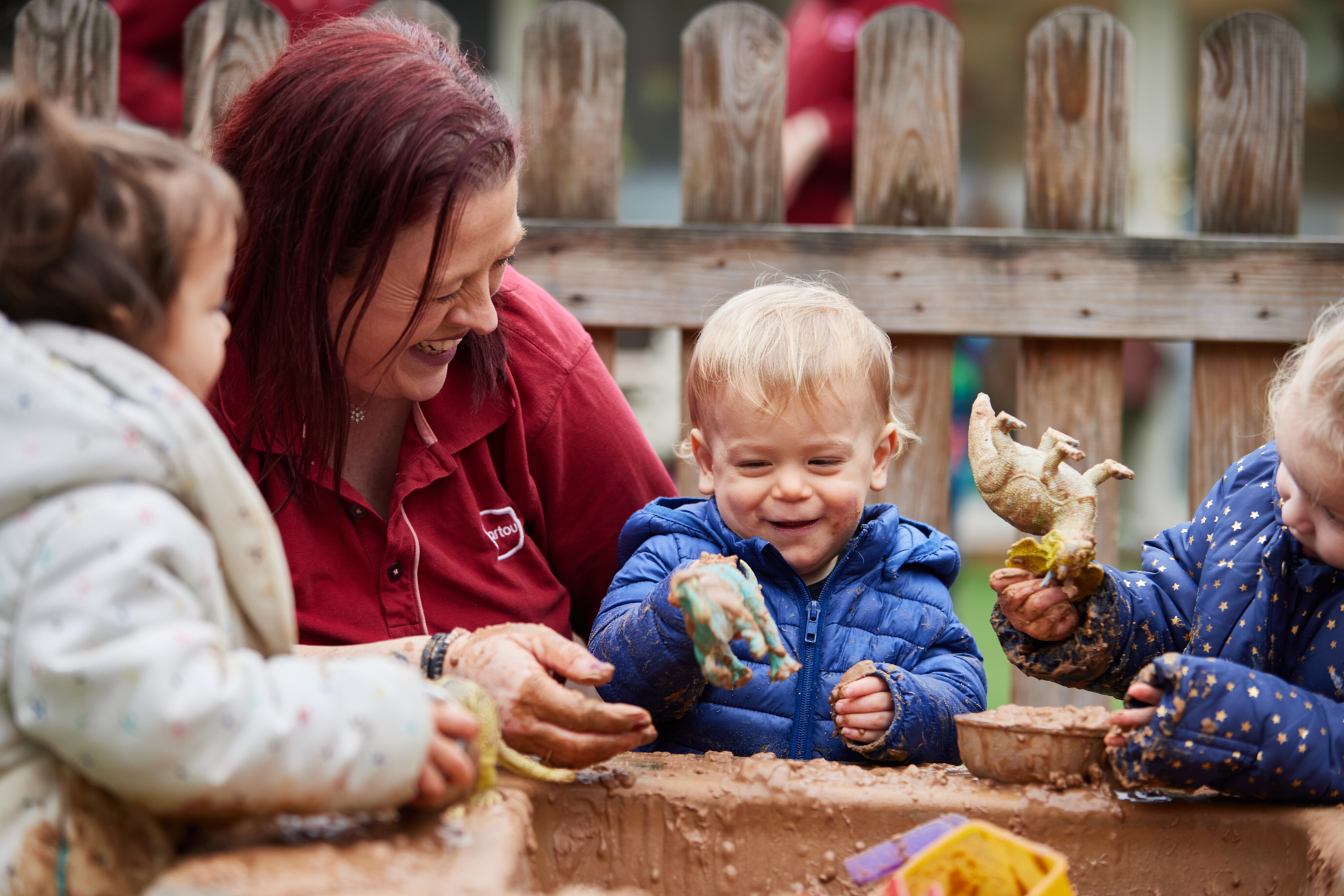 12:05Claude responded: A Partou practitioner in a red polo shirt plays alongside three toddlers at an outdoor mud kitchen, her hands fully covered in mud as she joins in with the chi…A Partou practitioner in a red polo shirt plays alongside three toddlers at an outdoor mud kitchen, her hands fully covered in mud as she joins in with the children