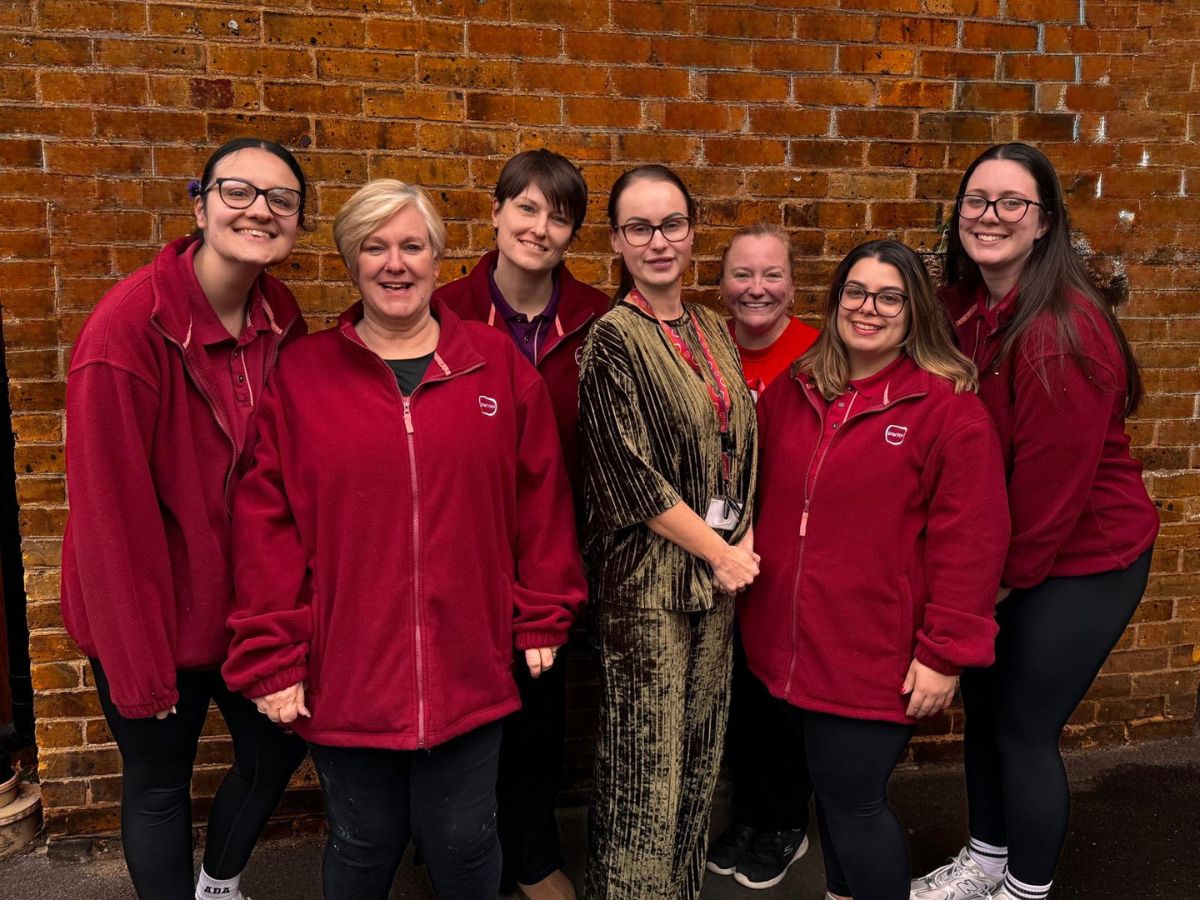A group of eight people standing close together in front of a red brick wall. Most are wearing matching red zip-up jackets and dark trousers, with some wearing glasses and white trainers. One person in the center is dressed differently in a patterned outfit with a lanyard. The group is arranged in two loose rows, leaning slightly toward the camera, and the scene appears to be outdoors.