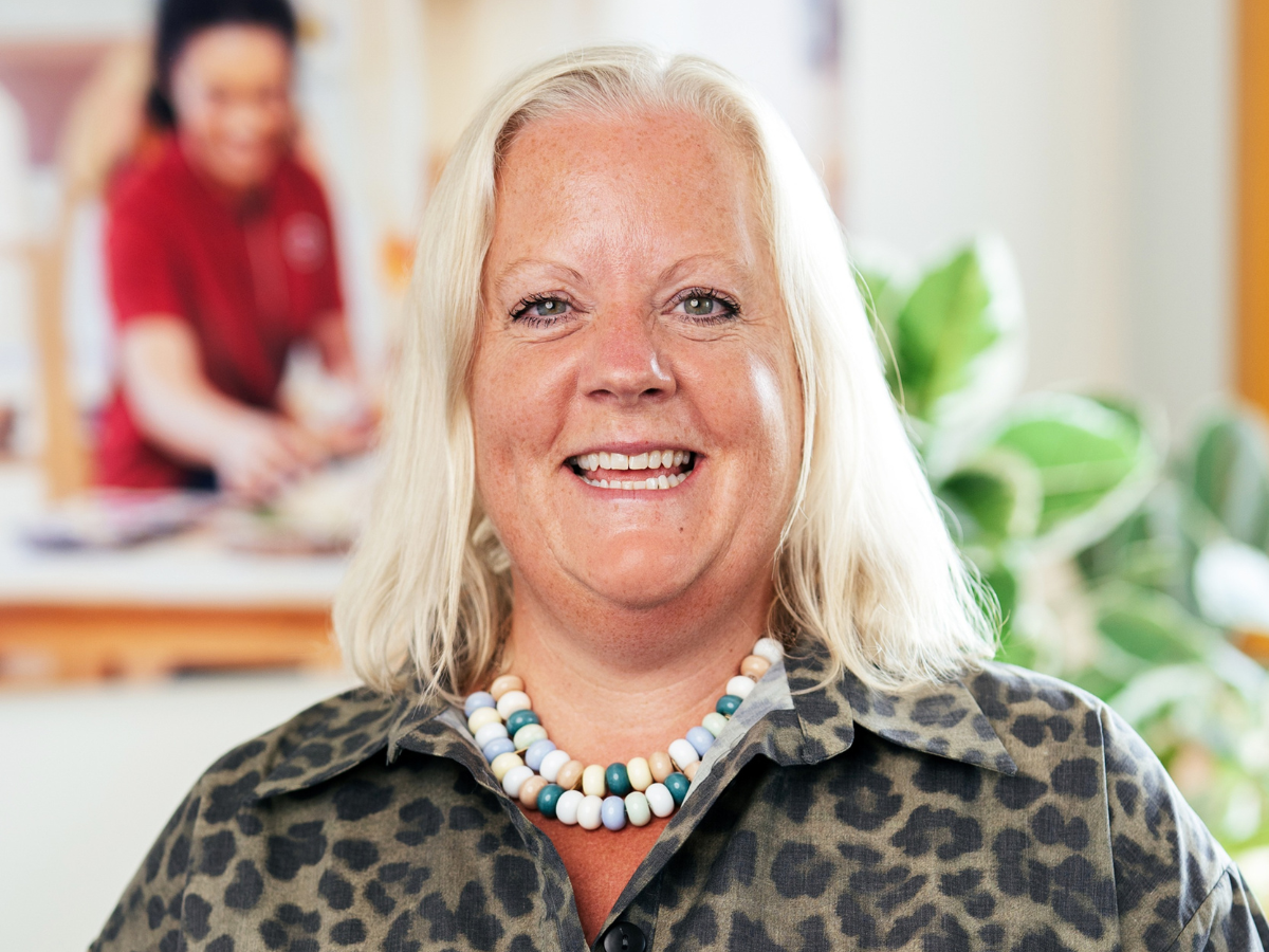 Head-and-shoulders portrait of Josie Mortimer, People & Culture Director at Partou, wearing a patterned button-up shirt and a colourful beaded necklace. Josie is facing the camera in a bright indoor setting. The softly blurred background shows a colleague in a red top and green houseplants, creating a warm and professional workplace environment.