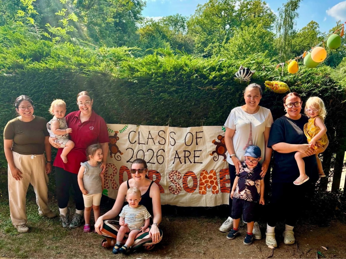 Group photo at Partou Bambinos Day Nursery & Pre-school in High Wycombe showing nursery staff, children and parents outdoors in front of a decorative banner reading “Class of 2026 Are Roarsome”. The celebration takes place in the nursery garden, with greenery, trees and colourful balloons visible in the background.