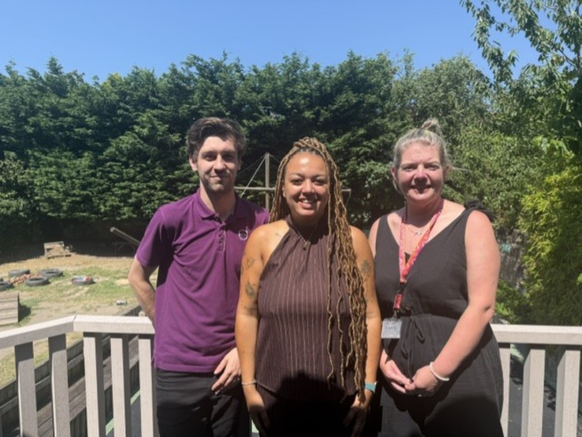 Three smiling nursery staff members from Partou Butterflies Day Nursery & Pre-school in Prenton, Birkenhead stand together outdoors in bright sunshine, posing on a wooden decking area with a garden and greenery behind them, celebrating a positive moment.